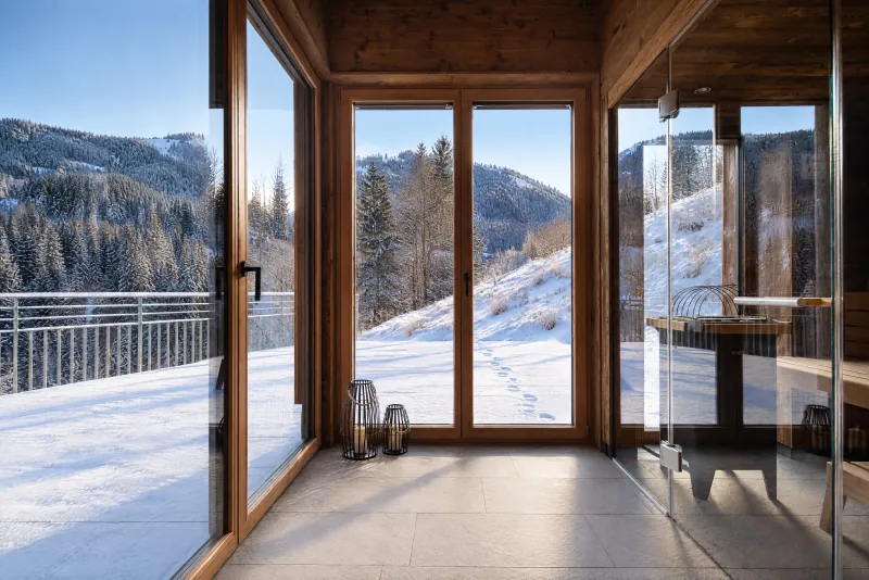 Blick durch raumhohe Fenster auf die verschneite Terrasse und die Berge