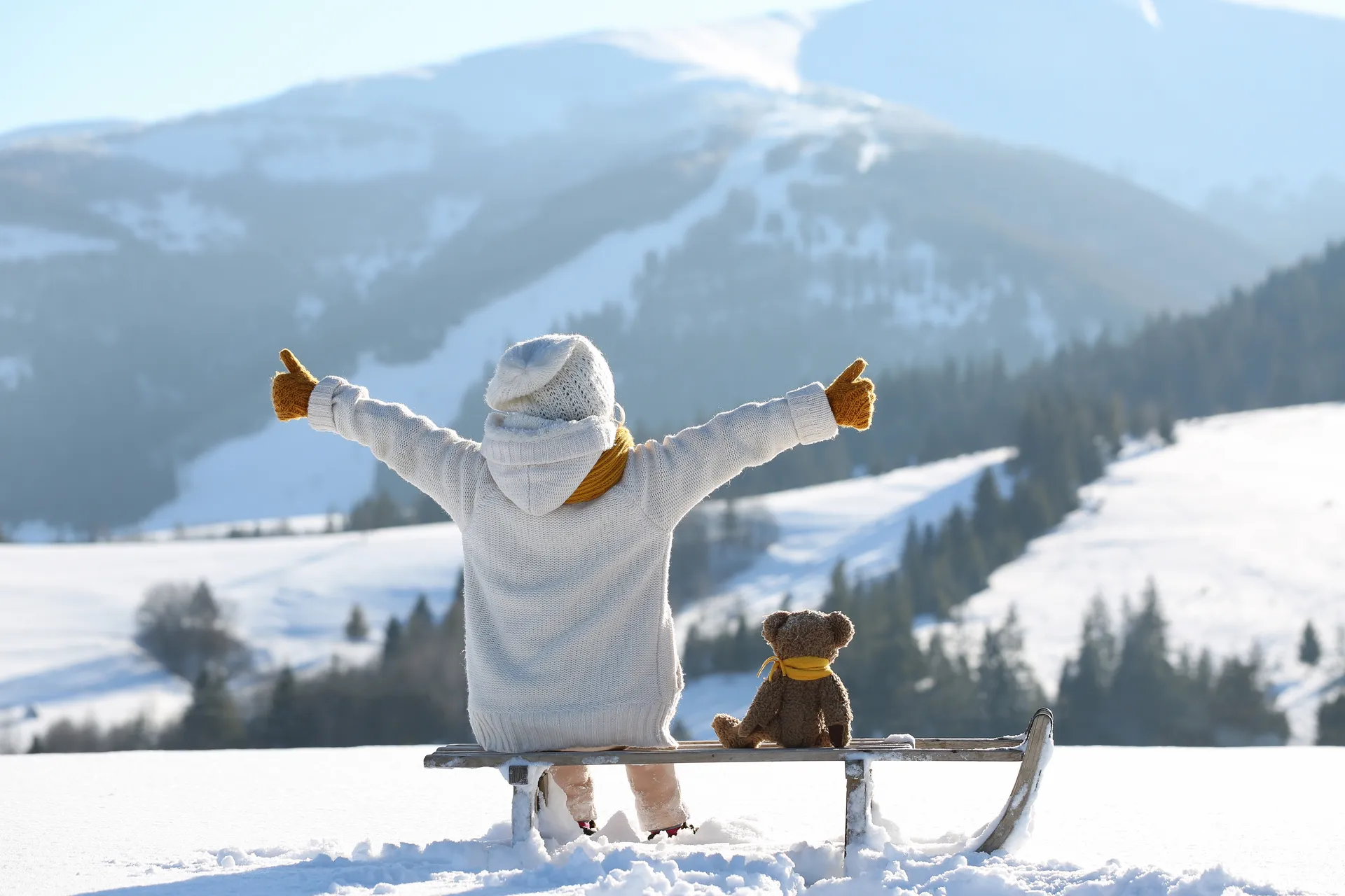 Kind mit Teddybär auf dem Schlitten blickt auf die verschneiten Berge