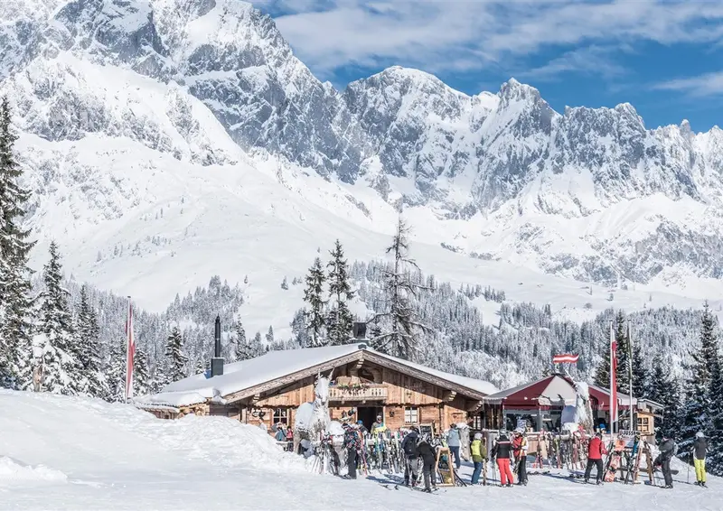 Verschneite Berghütte mit Skifahrern vor Bergpanorama